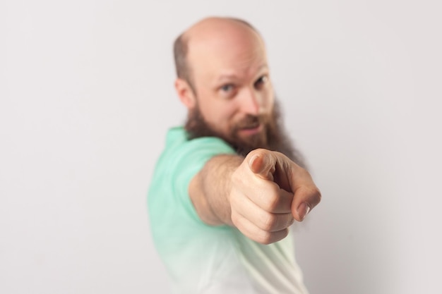 Portrait of funny middle aged bald man with long beard in light green t-shirt standing, pointing and looking at camera. focus on finger. indoor studio shot, isolated on grey background.