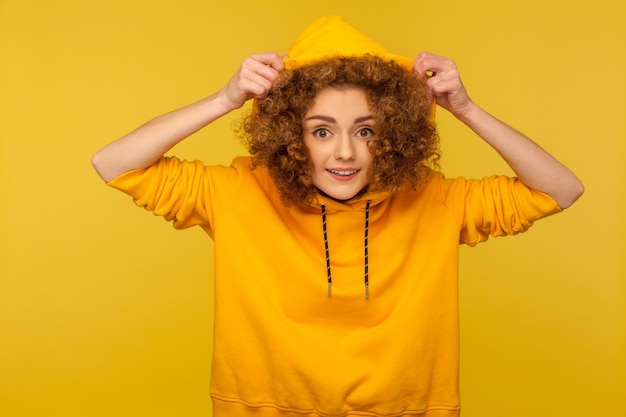 Portrait of funny positive girl with curly hair wearing urban style hoodie, putting on hood and smiling at camera, youth sport style female fashion. indoor studio shot isolated on yellow background