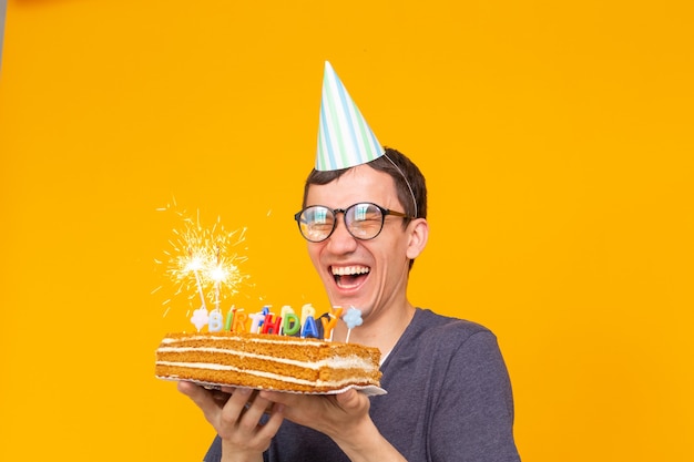 Ritratto di un ragazzo divertente e positivo con un tappo di carta e occhiali che tengono una torta di congratulazioni nelle sue mani su una superficie gialla