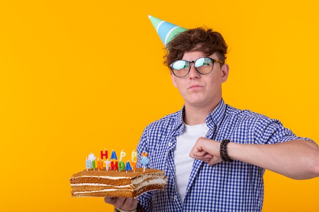 Ritratto di un ragazzo divertente e positivo con un berretto di carta e bicchieri che tengono una torta di congratulazioni nella sua