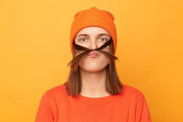 Портрет забавной позитивной женщины-хипстера в оранжевом свитере и шляпе, позирующей изолированно на желтом фоне, делающей усы с волосами и посылающей воздушный поцелуй