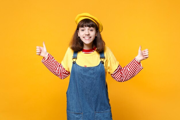 Portrait of funny pretty girl teenager in french beret, denim sundress showing thumbs up isolated on yellow wall background in studio. people sincere emotions, lifestyle concept. mock up copy space