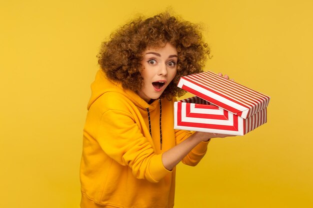 portrait of funny shocked curlyhaired woman in hoodie looking at camera with amazed expression and holding opened gift box surprised by birthday present studio shot isolated on yellow background