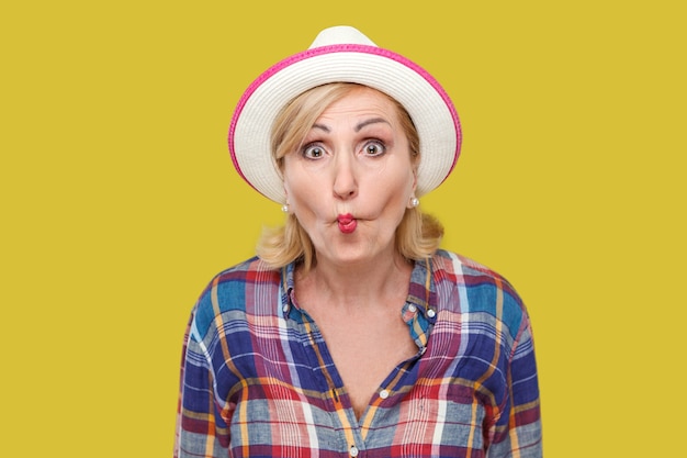 Portrait of funny shocked modern stylish mature woman in casual style with white hat standing with fish lips and looking at camera with crazy face. indoor studio shot isolated on yellow background.