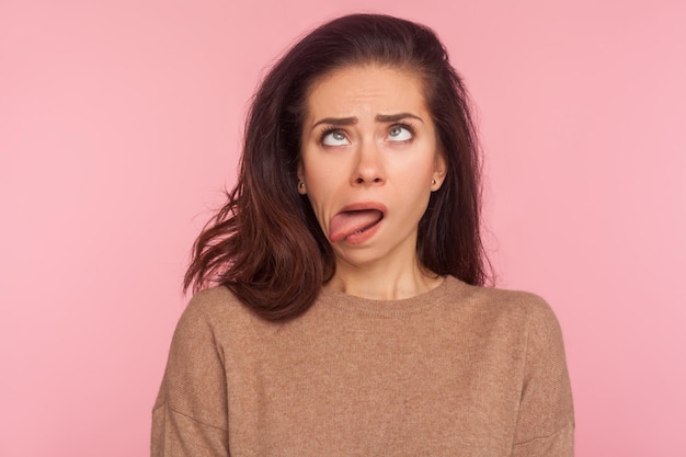 Portrait of funny silly young woman with brunette hair looking cross eyed and sticking tongue out, making dumb brainless face, disobedient goofy expression. studio shot isolated on pink background