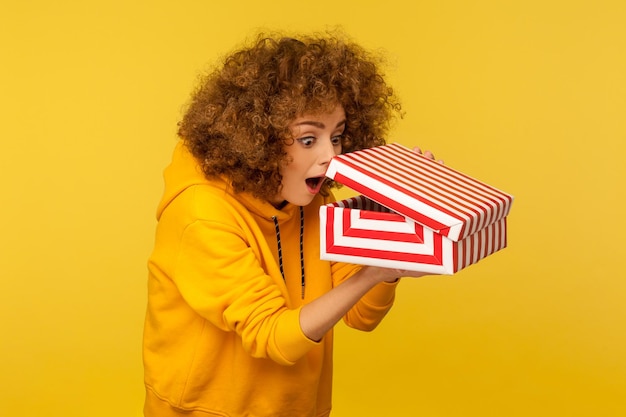 Portrait of funny surprised curious curlyhaired woman in hoodie looking into gift box unwrapping present and peeking inside with interest open mouth in amazement indoor studio shot isolated