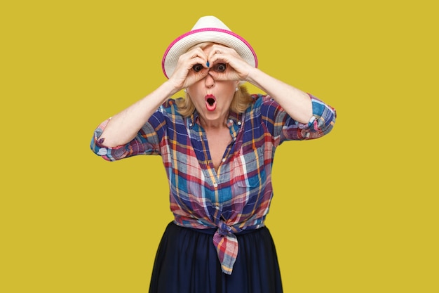 Photo portrait of funny surprised modern stylish mature woman in casual style with hat standing with binoculars gesture and looking with amazed face. indoor studio shot isolated on yellow background.