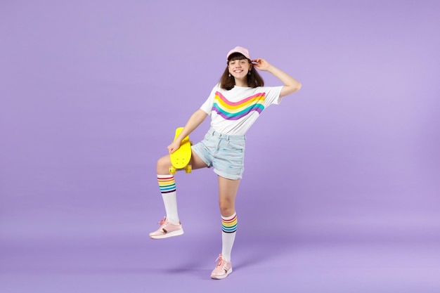 Portrait of funny teen girl in vivid clothes standing, holding yellow skateboard isolated on violet pastel wall background in studio. People sincere emotions, lifestyle concept. Mock up copy space.