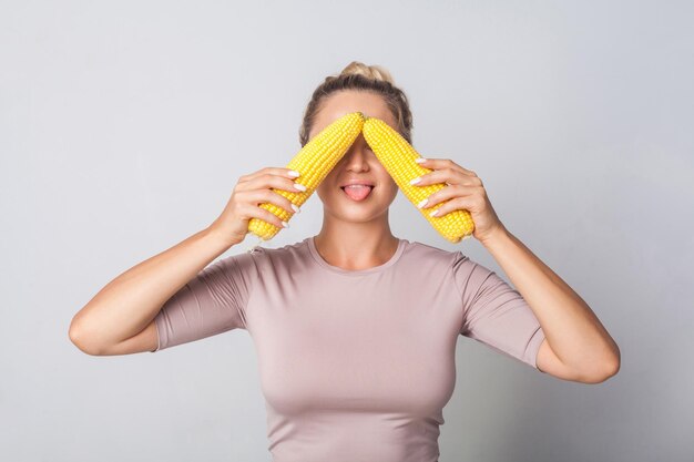 Portrait of funny woman covering eyes with corn cobs and sticking out tongue concept of healthy eating and vegetarian diet fresh raw vegetables low calorie food indoor studio shot grey background