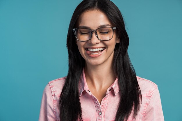 Portrait of funny woman wearing glasses fooling around, showing tongue out and closed her eyes with comical expression. Indoor studio shot isolated on blue background