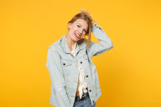 Portrait of funny young woman in denim casual clothes looking up, putting hand on head, showing tongue isolated on bright yellow orange wall background. People lifestyle concept. Mock up copy space.