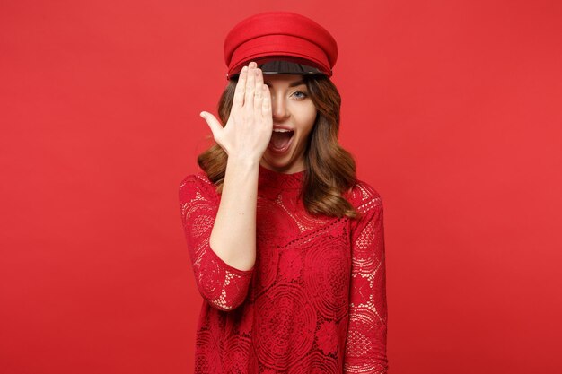 Portrait of funny young woman in lace dress cap keeping mouth open, covering face with palm isolated on bright red background in studio. People sincere emotions, lifestyle concept. Mock up copy space.