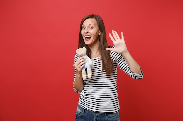 Portrait of funny young woman in striped clothes holding teddy bear plush toy showing palm, waving hand 