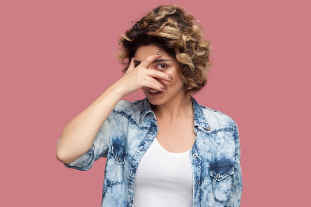 Portrait of funny young woman with curly hairstyle in casual blue shirt standing, covering her eyes and looking through fingers. spy or shy concept. indoor studio shot, isolated on pink background.