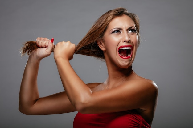 Portrait of a furious young woman in red dress pulling her hair and shouting.