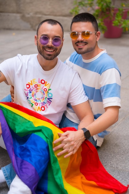 Photo portrait of gay male couple sitting on the floor smiling at pride party with rainbow flag lgbt concept