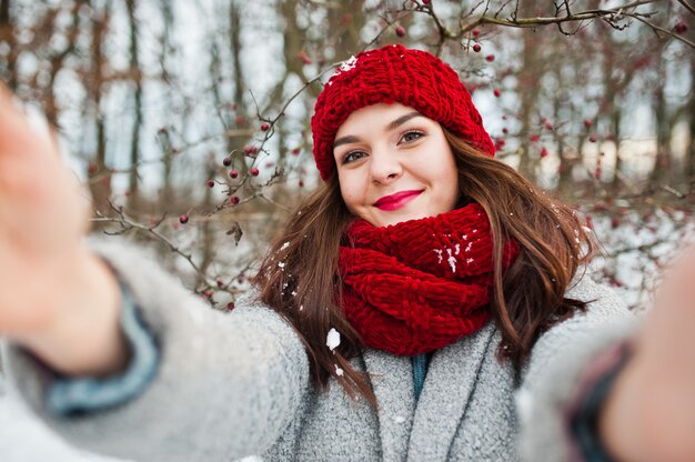 Portrait of gentle girl in gray coat , red hat and scarf near the branches of a snow-covered tree