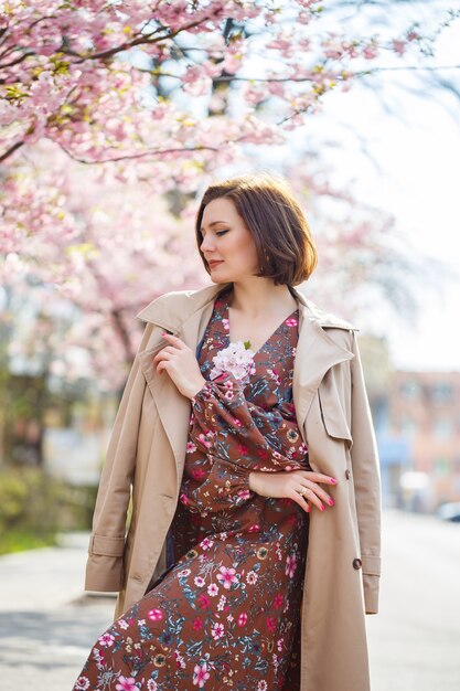 Portrait of a gentle woman against the background of sakura flowers. Walk in the blossoming sakura garden. Young stylish woman standing in sakura park and enjoying beauty