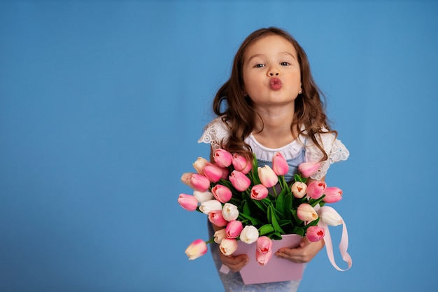 Portrait of a girl on a blue background who holds a bouquet with tulips and blows a kiss.