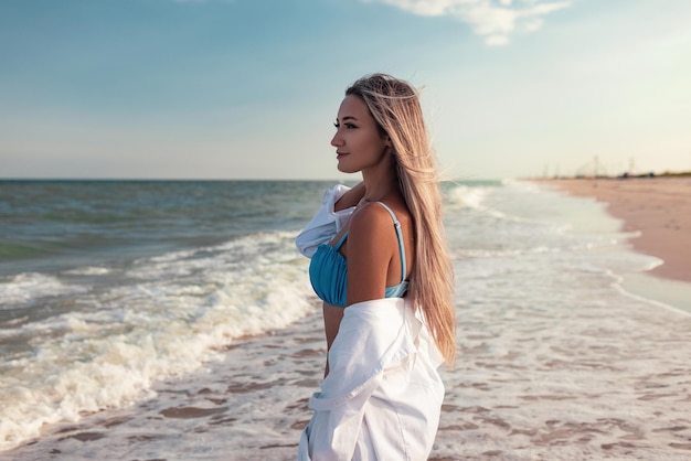 Ritratto di una ragazza in costume da bagno blu e camicia bianca sullo sfondo del mare blu e del cielo limpido