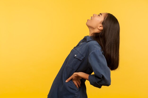 Photo portrait of girl in denim shirt shouting from acute low back pain, suffering kidney stone, hips joint inflammation, pinched nerve or spinal injury. indoor studio shot isolated on yellow background