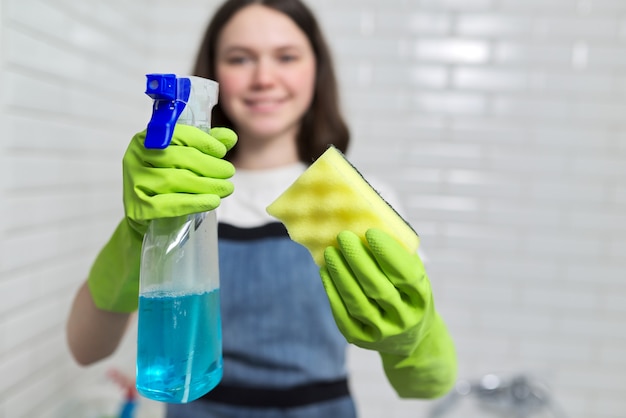 Portrait of girl doing cleaning in bathroom. Teenager in apron gloves with detergent and rag sponge smiles looking at camera. Housekeeping, cleanliness at home, service, young people concept