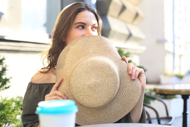 여름 거리 카페에 앉아 있는 동안 밀짚 모자 뒤에 얼굴을 숨기고 있는 소녀의 초상화.