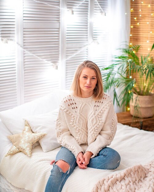 Portrait of a girl at home dressed in a warm knitted sweater, sitting in jeans on the bed. bright bedroom with plants