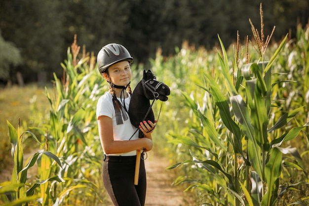 A portrait of a girl a jockey in a helmet and a white Tshirt who holds a black horse in her hands a toy on a stick stands in the corn Hobbyhorsing