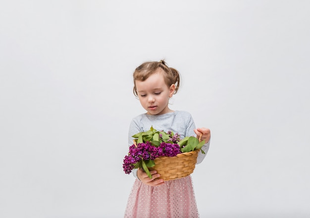 Portrait of a girl looks at the basket of lilacs. Cute girl in a grey dress with a tail on a white space.Copy space