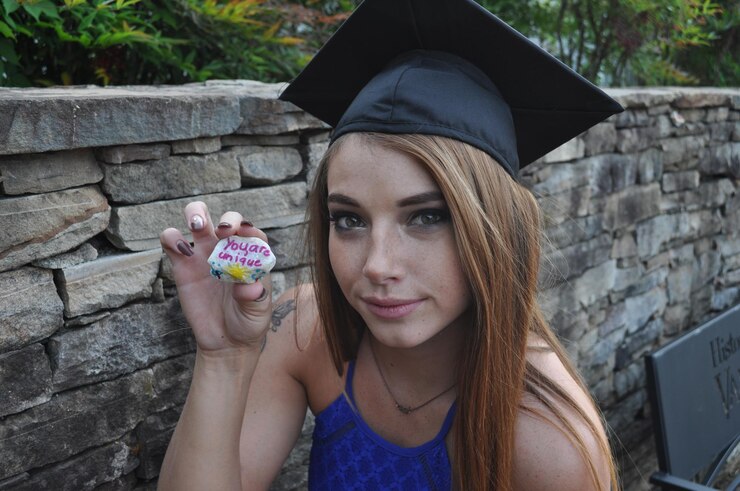 Photo portrait of girl in mortarboard holding stone with text by wall