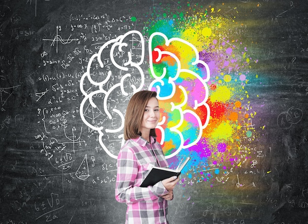 Portrait of a girl in a pink checkered shirt standing with an open book near a blackboard with a large and colorful brain sketch.