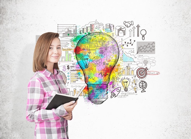Portrait of a girl in a pink checkered shirt standing with an open book near a concrete wall with a large colorful light bulb sketch and business icons.