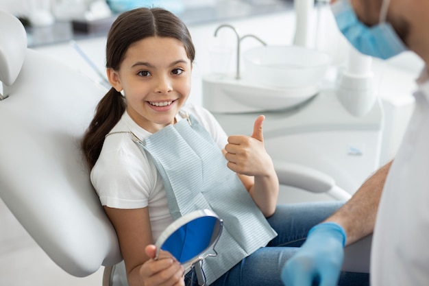 Portrait of girl sitting in dental chair showing thumb up and smiling at the camera