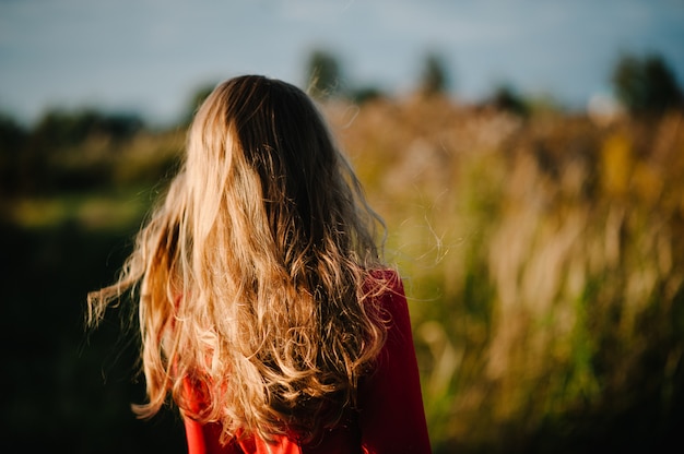 Ritratto di una ragazza in piedi indietro in autunno in un abito rosso contro il campo sulla natura. la metà superiore. avvicinamento. vista dal retro.