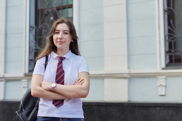 Portrait of girl student of 17, 18 years old in white T-shirt with backpack, female looking at camera, outdoor, building background, copy space