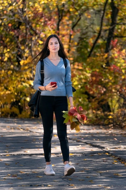 秋の公園の路地を歩いている女子学生の肖像画。手に電話を持つ若い美しい女性。縦フレーム