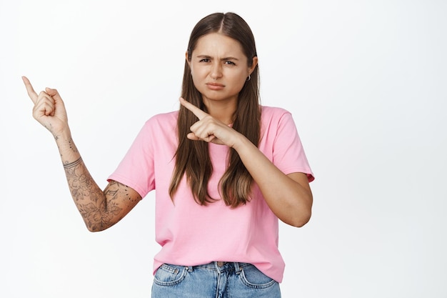 Portrait of girl with doubts, pointing at upper left corner and squinting, frowning skeptical, being displeased, standing over white background