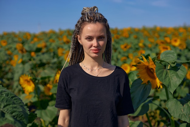 Ritratto di una ragazza con i dreadlocks che sta in mezzo a un luminoso campo di girasoli e guarda...