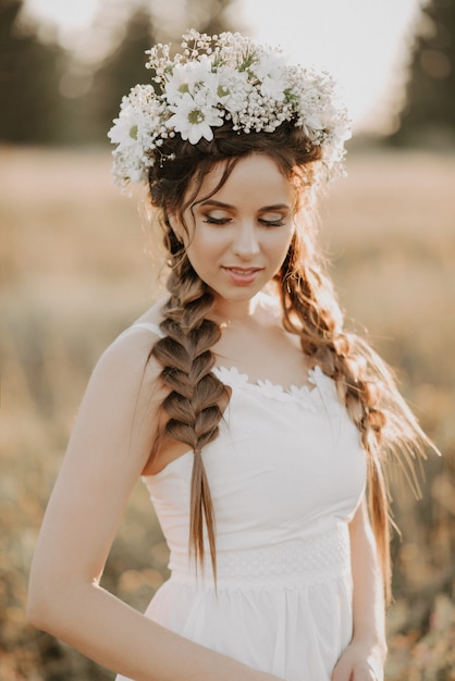 Portrait of girl with flower wreath and braids in white dress in summer field at sunset