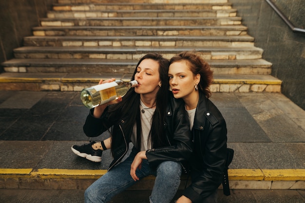 Portrait girls in leather jackets and jeans drinking wine from a bottle on the stairs