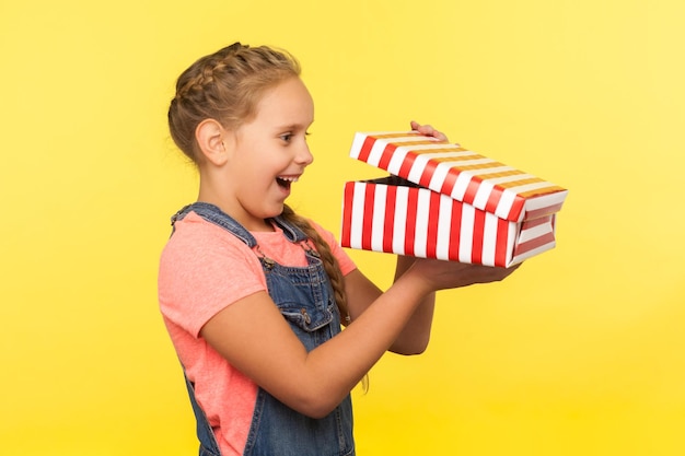 Portrait of glad curious little girl with braid in denim overalls peeking into gift box and laughing from happiness, enjoying best surprise, holiday bonus. studio shot isolated on yellow background