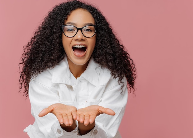 Portrait of glad dark skinned woman keeps palms outstretched friendly expression sends air kiss to lover has curly hairstyle wears optical eyewear and white jacket poses against pink background