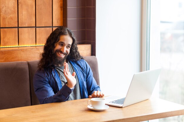 Portrait of glad handsome young adult man freelancer in casual style sitting in cafe and talking with his friend in laptop showing goodbye gesture with palm Indoor lifestyle concept