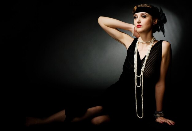 Portrait of a glamorous dark haired model with perfect makeup wearing a luxurious necklace black bodysuit and long top holding her hands behind her head and looking straight ahead