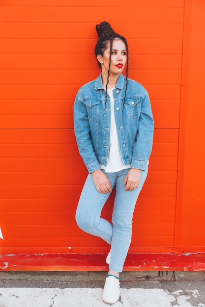 Foto ritratto di una donna affascinante con i dreadlocks in un abito di jeans e con le labbra rosse contro un muro arancione