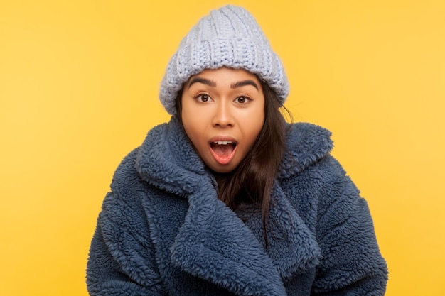 Portrait of glamour fashion model in stylish urban outfit, fur coat and winter hat, standing with open mouth, looking with surprised shocked expression. studio shot isolated on yellow background