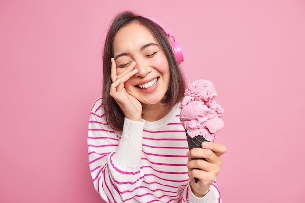 Portrait of good looking brunette Asian woman keeps hand on face eyes closed smiles broadly enjoys spare time holds tasty appetizing ice cream dressed in striped jumper isolated over pink wall