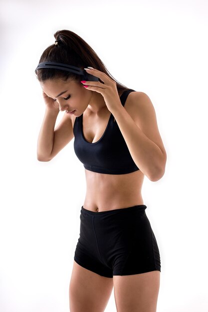 Portrait of good-looking caucasian woman in black top and shorts listening to music in headphones during sport, going to do workout, looking down, isolated on white studio background