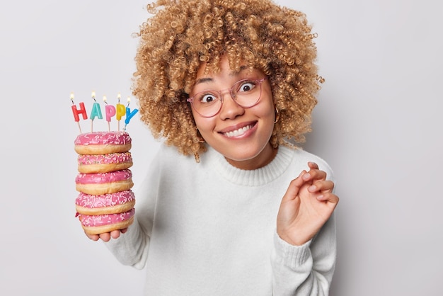 Portrait of good looking curly woman bites lips looks with\
curious expression holds pile of glazed doughnuts with burning\
candles wears spectacles casual jumper isolated over white\
background